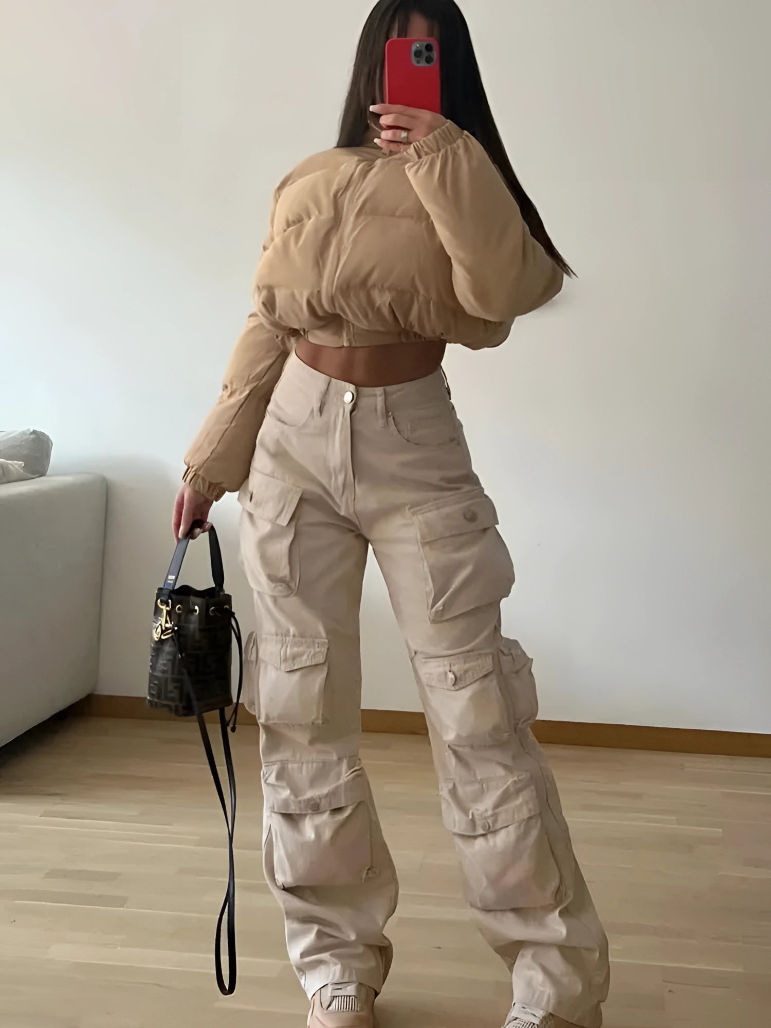 A model wearing beige cargo pants paired with a cropped puffer jacket, striking a trendy street style pose with one hand on the hip, standing near a couch with a bag on the floor.