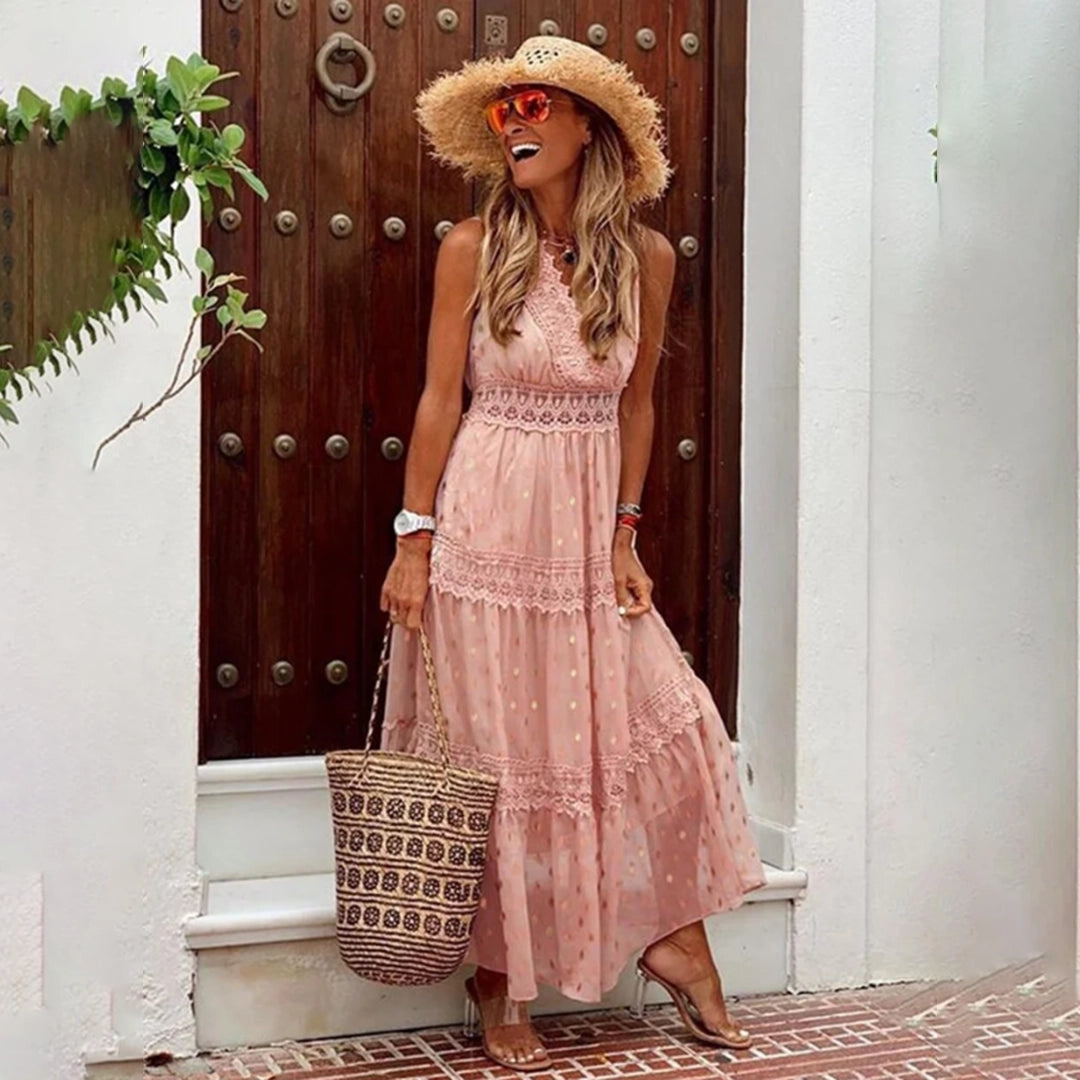 A woman wearing a sleeveless, lightweight beige lace bohemian maxi dress, standing outdoors next to a wooden structure, holding a hat. The dress is perfect for summer wear and is available in pink and 2XL.