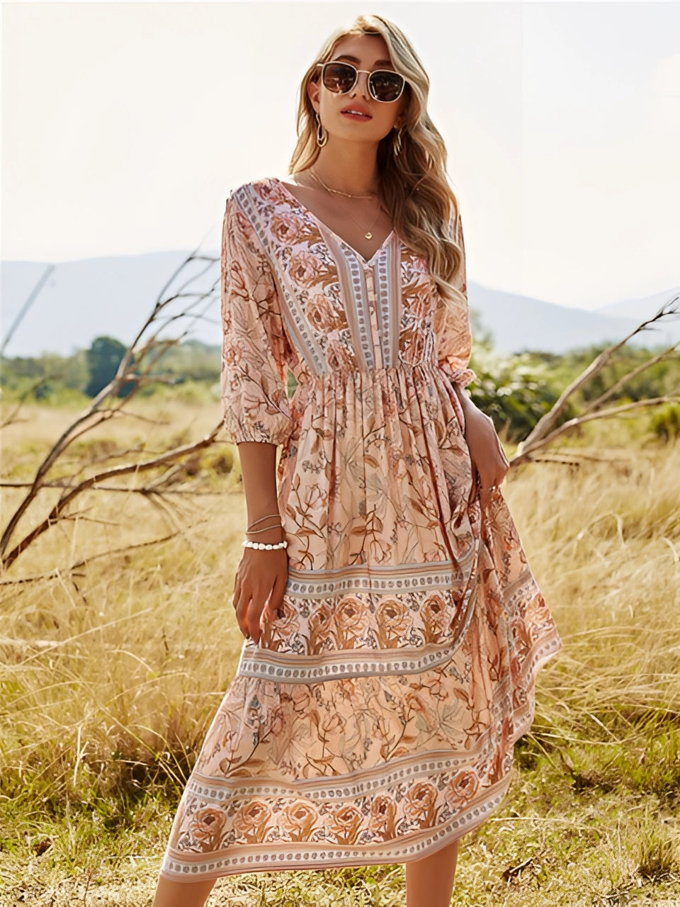 A woman wearing a blue floral Bohemian maxi dress with a V-neckline, standing in a grassy area under a clear sky, smiling and looking happy.