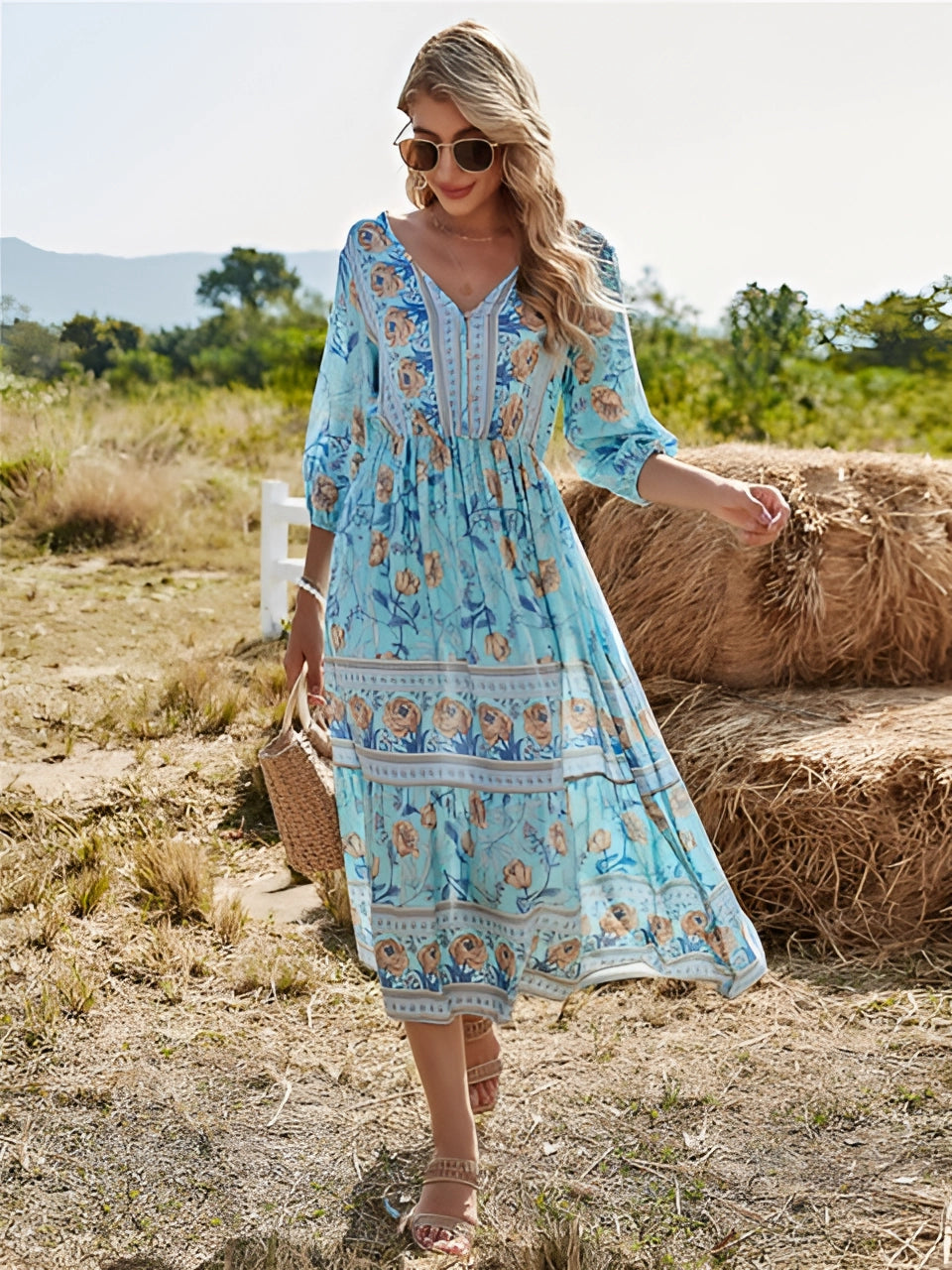 A woman wearing a blue floral Bohemian maxi dress with a V-neck, standing outdoors in nature. She has sunglasses on her head and is surrounded by plants.