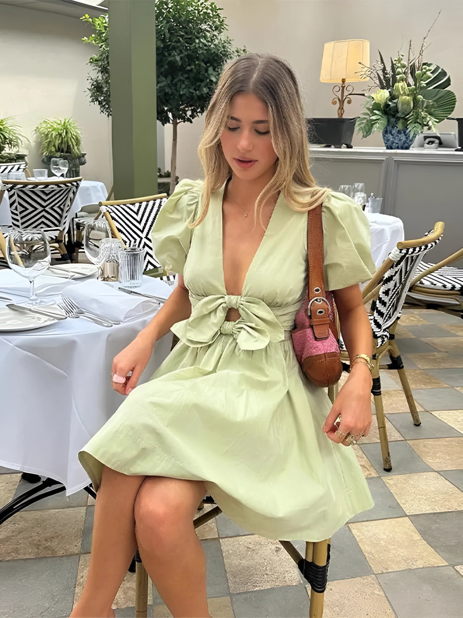 Light green puffy sleeve mini dress with bow waist worn by a model, standing beside a chair and a plant.